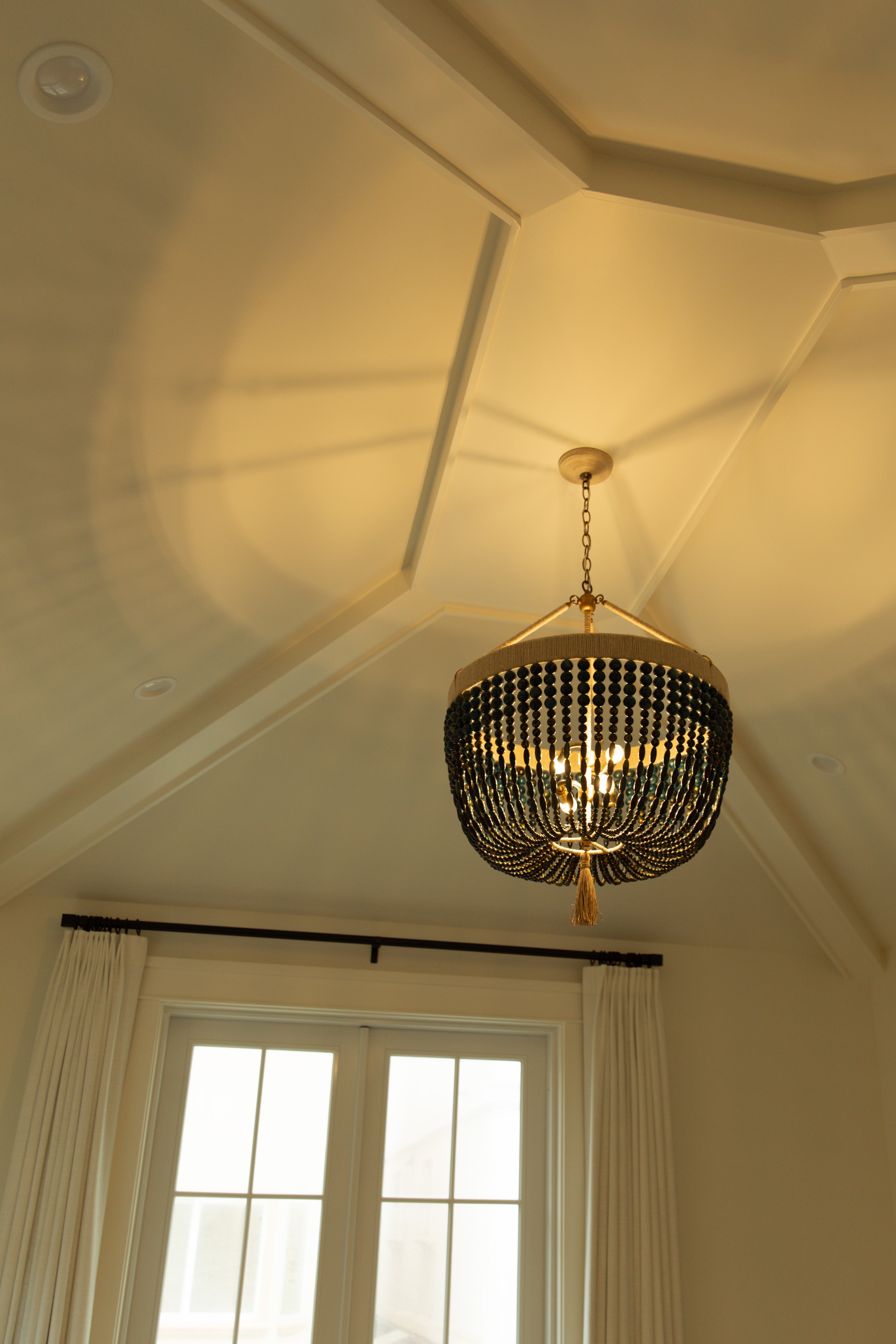 Looking up at a striking dark beaded chandelier hanging from a custom vaulted ceiling with geometric trim details in a light-filled primary bedroom.