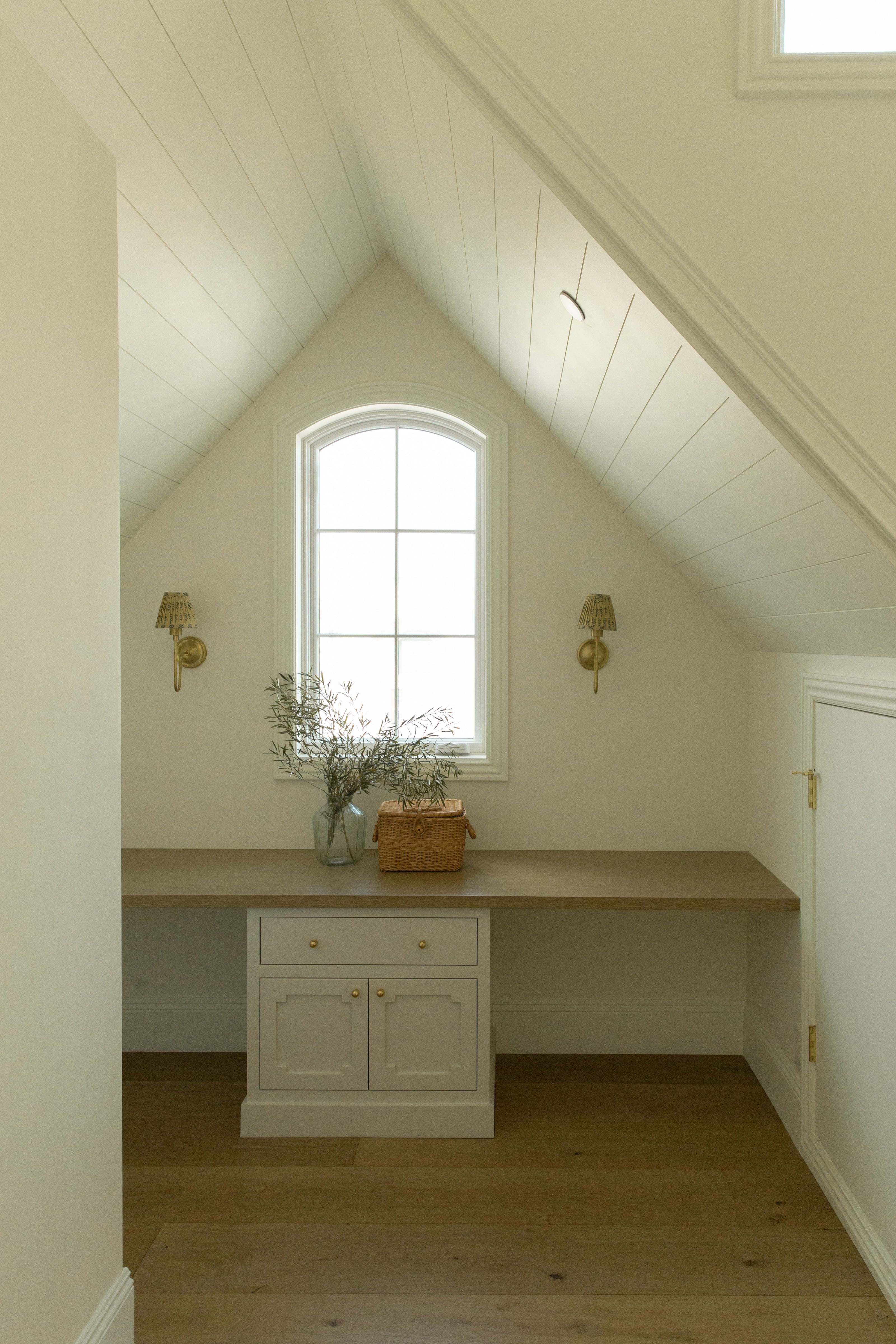 Custom home office featuring a built-in wood desk, an arched window, brass sconces, and a white paneled vaulted ceiling designed by a Seal Beach interior designer.