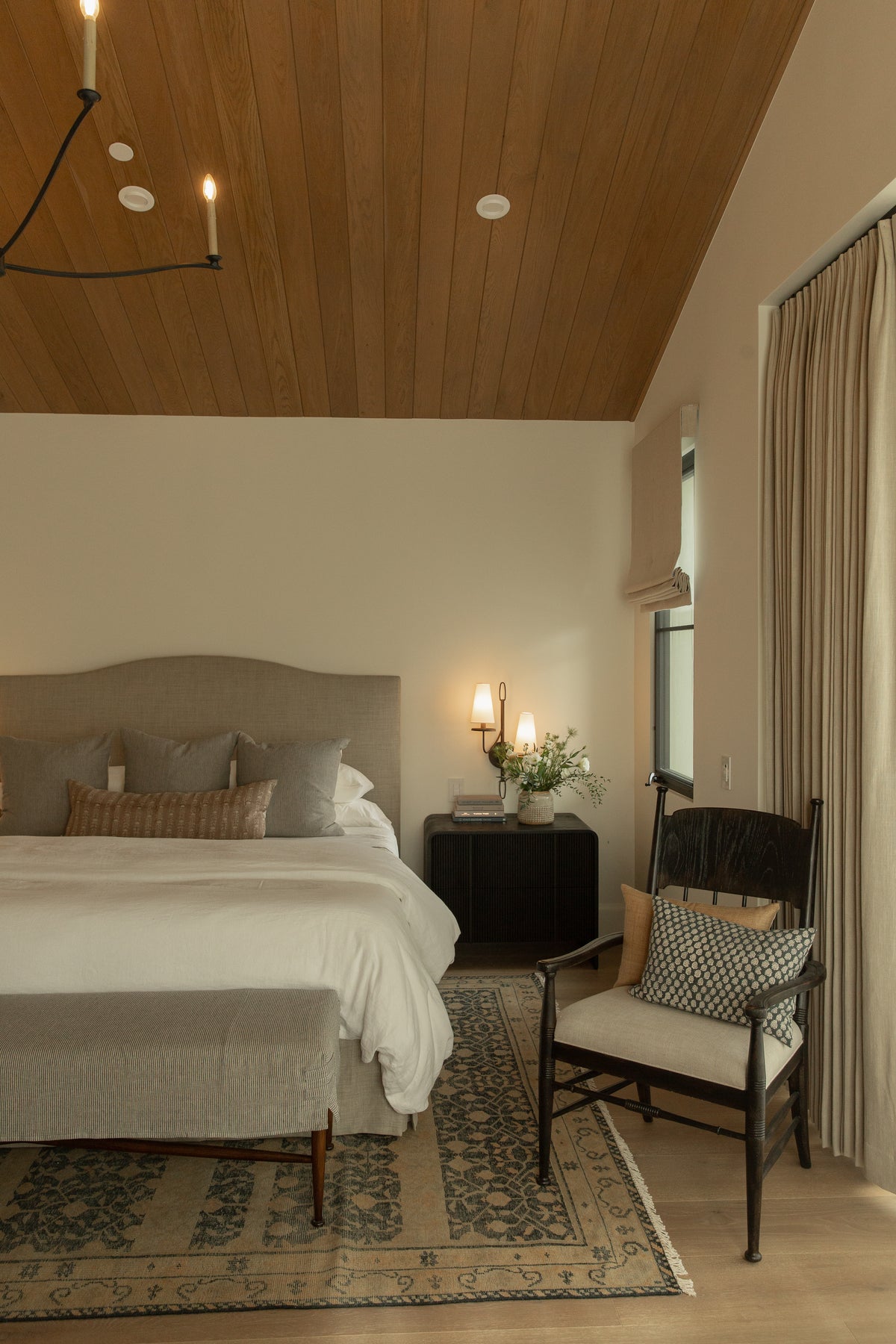 A beautifully styled transitional bedroom highlighting a rich wood-paneled ceiling, a sleek dark iron chandelier, an upholstered headboard, and a vintage woven chair resting on a patterned area rug.