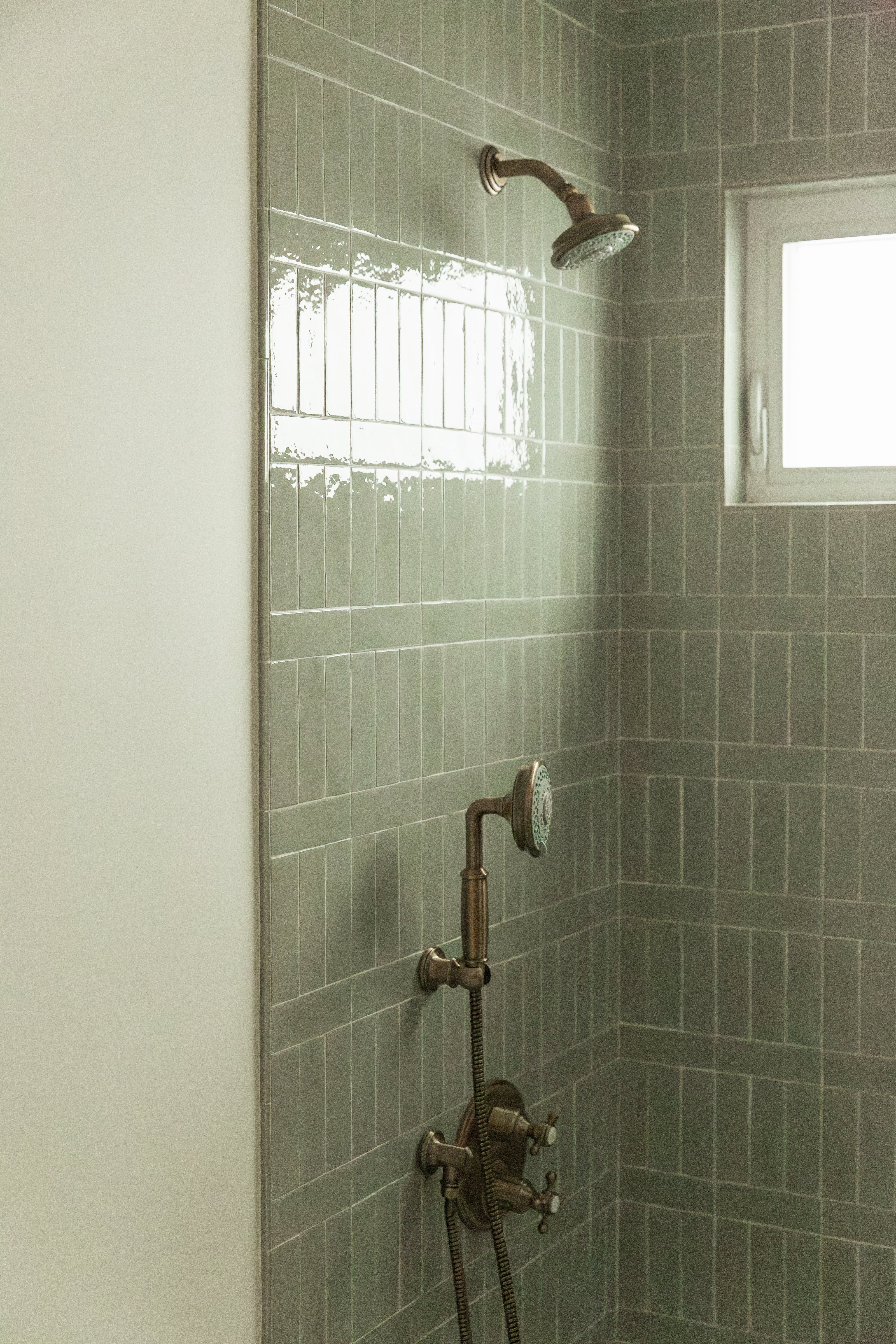 Close-up of a custom shower design featuring vertically stacked glossy sage green subway tile with a horizontal accent band, beautifully paired with vintage-style antique brass plumbing fixtures including a showerhead, hand wand, and cross-handle temperature controls.