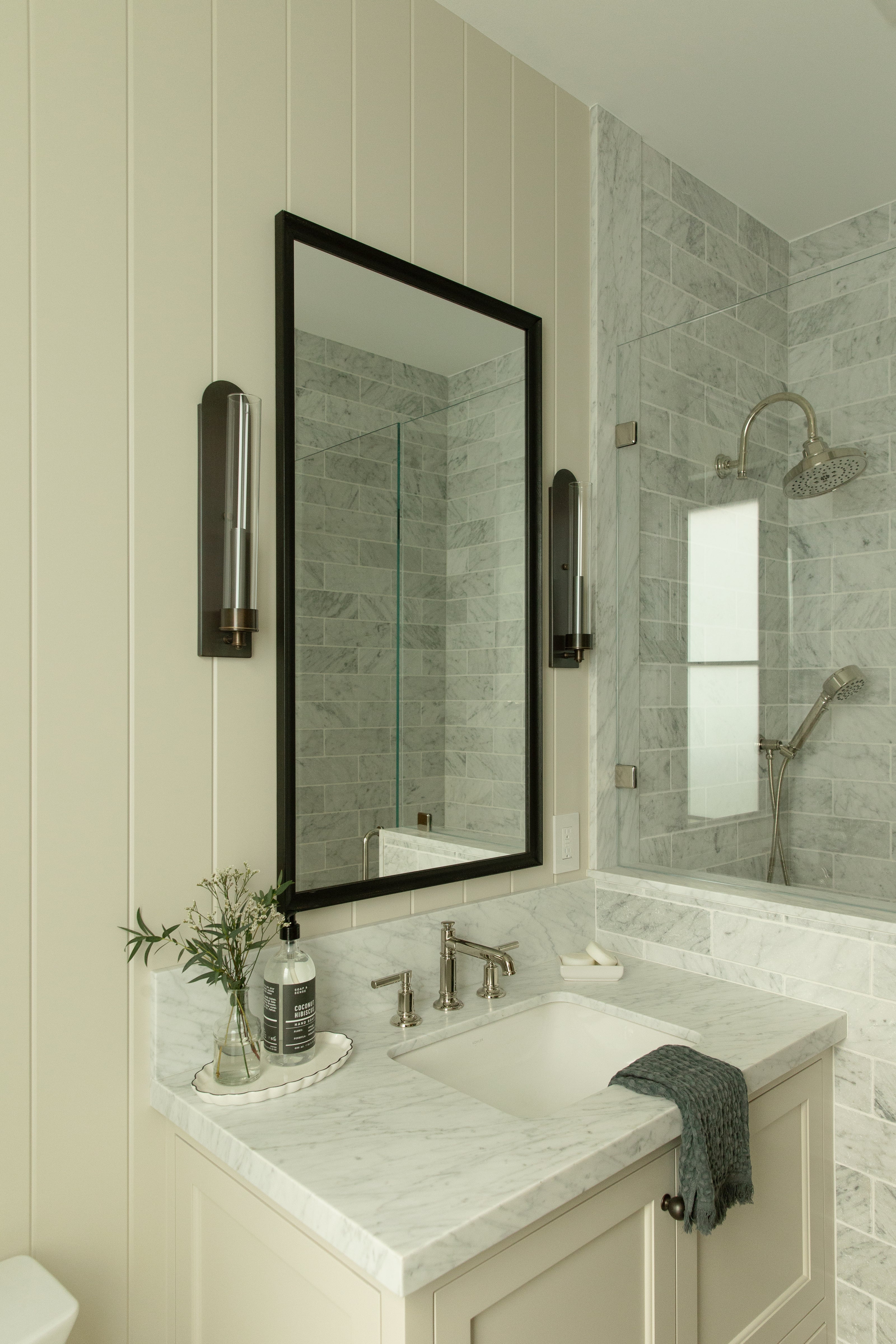 A bright transitional guest bathroom featuring cream vertical shiplap walls, a black framed mirror flanked by tall glass sconces, and a marble vanity countertop next to a glass-enclosed shower.
