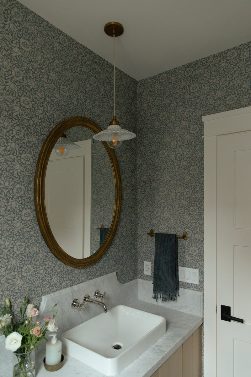 A coastal farmhouse powder room featuring a fluted glass pendant light, an oval brass mirror, a wall-mounted faucet, and a classic white paneled door.