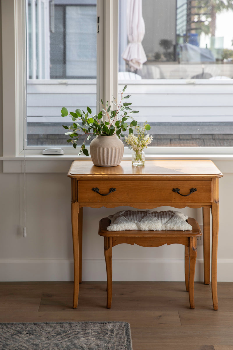 A charming vintage wood writing desk styled with a textured ceramic vase, fresh greenery, and a matching small stool with a plush cushion.