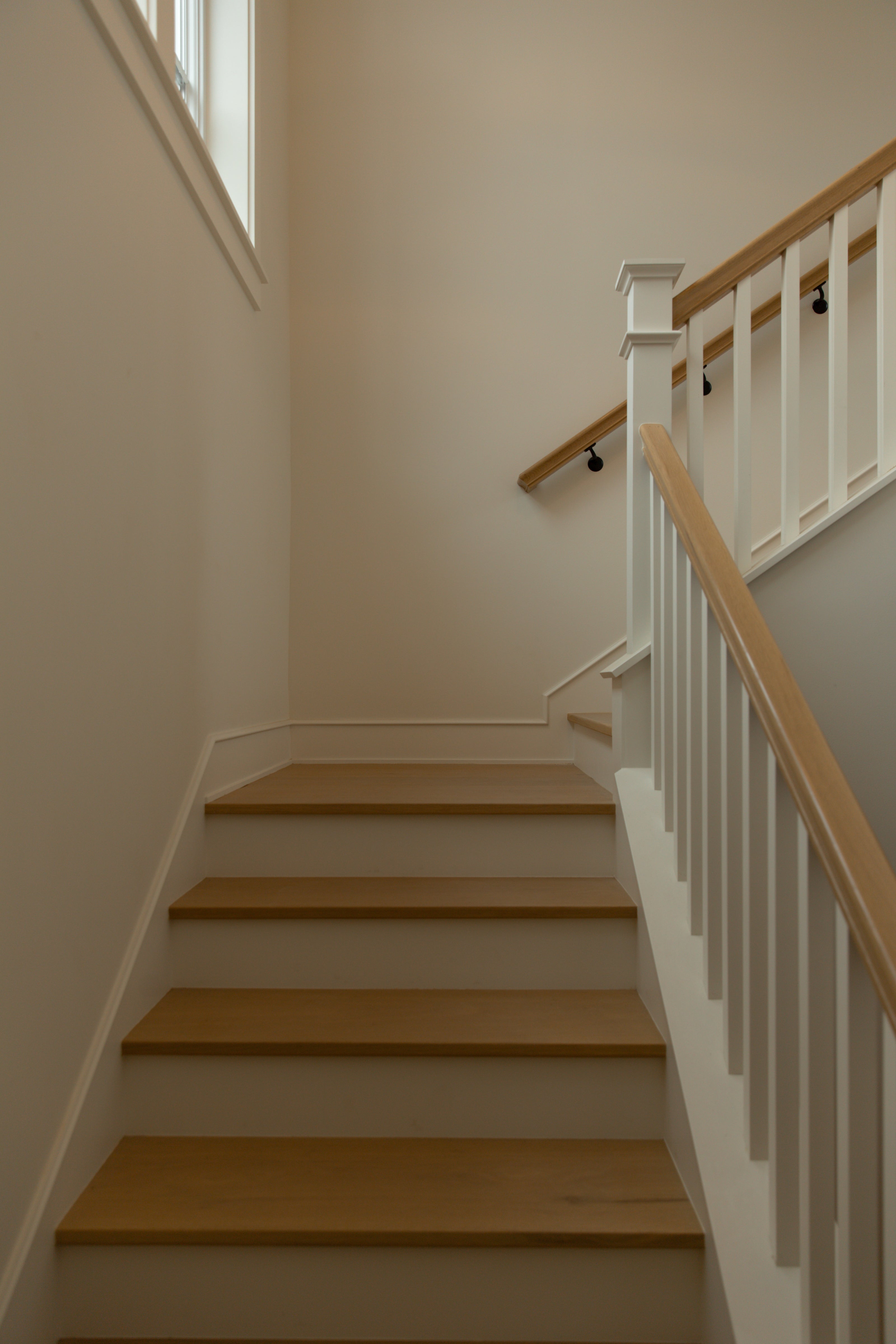 A classic coastal farmhouse staircase design showcasing light white oak stair treads beautifully contrasted against crisp white painted risers and black wall-mounted handrail brackets.