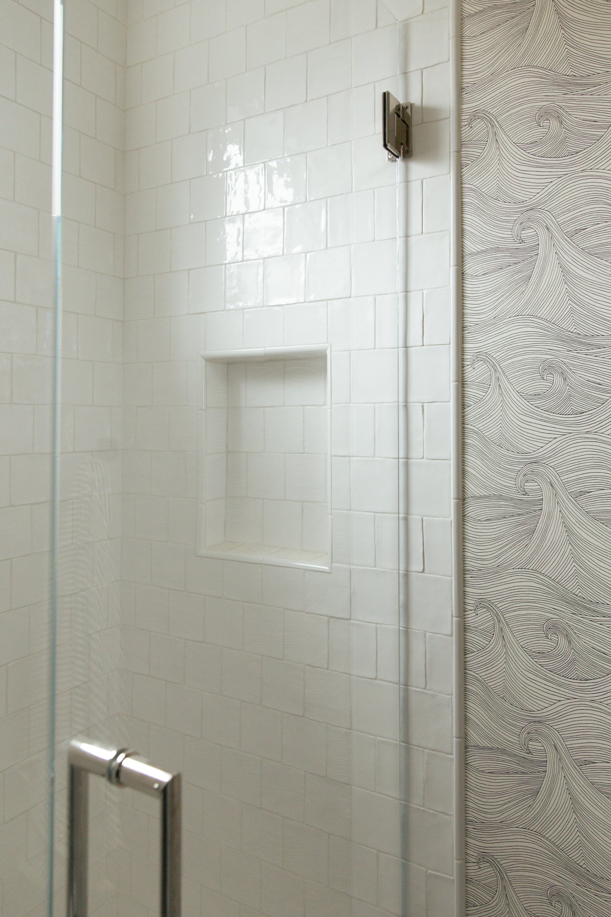 A seamless glass walk-in shower finished with glossy white square tiles and a built-in shower niche, contrasting perfectly with the adjacent wave patterned bathroom wallpaper.