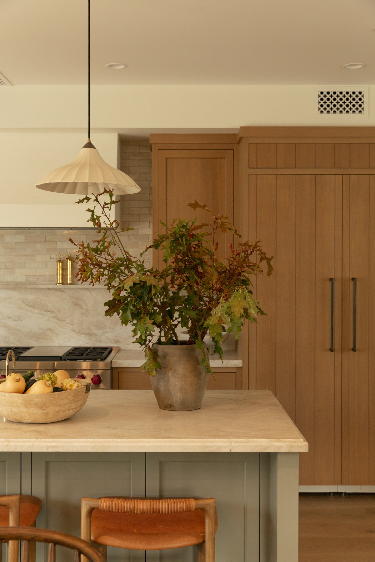 Detail of a kitchen island styled with a large rustic vase and fall foliage, showcasing woven leather and teak counter stools alongside a floor-to-ceiling light oak panel-ready refrigerator.