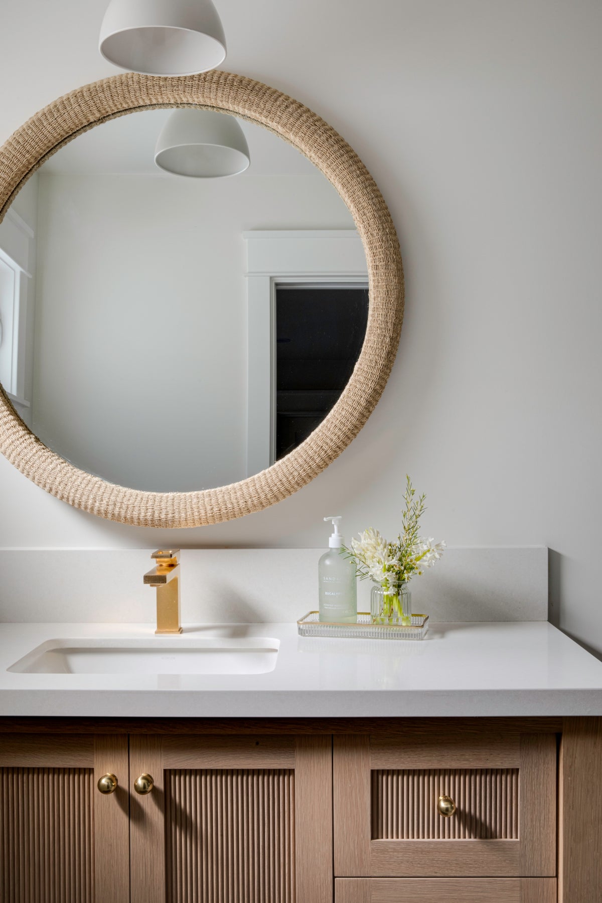 A coastal-inspired bathroom vanity featuring a round woven rope mirror, a white quartz countertop, and custom light oak fluted cabinetry.